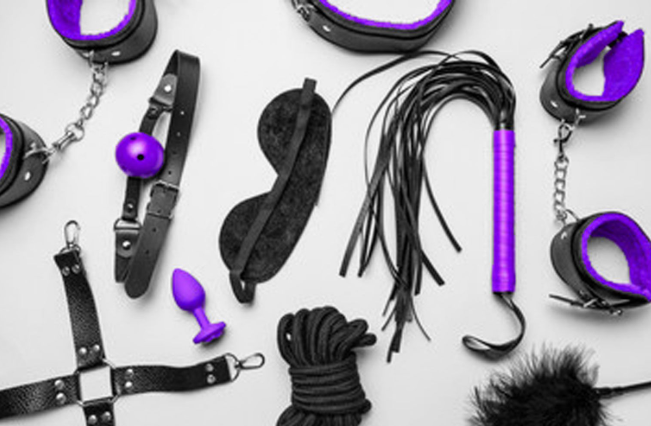 Black and purple BDSM accessories, including cuffs, a blindfold, rope, a flogger, and restraints, arranged on a light background